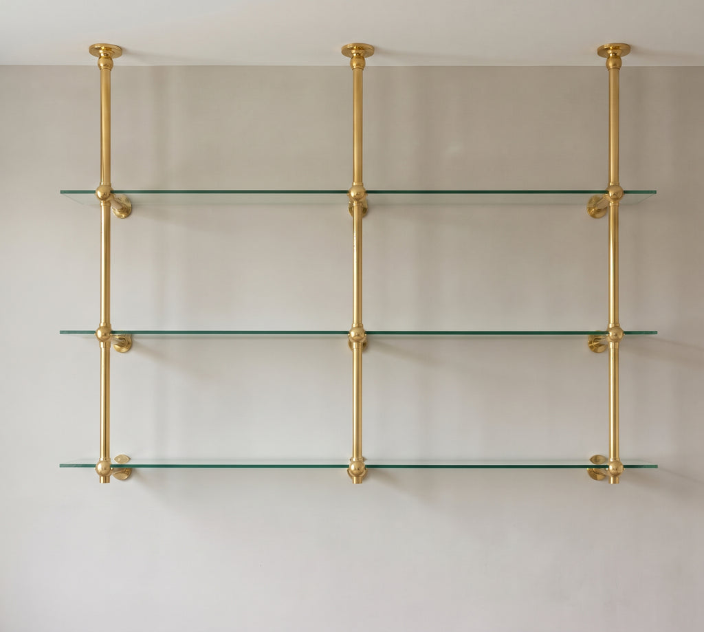 Front view of the two-bay brass bistro shelf with three ceiling-mounted posts and three clear glass shelves