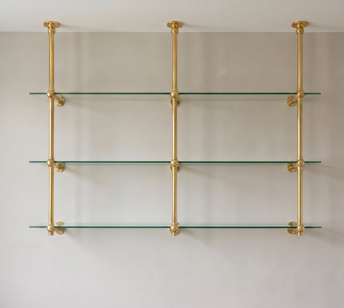 Front view of the two-bay brass bistro shelf with three ceiling-mounted posts and three clear glass shelves
