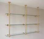 Angled view of the unlacquered brass shelf: three ceiling posts with wall arms holding three tiers of glass