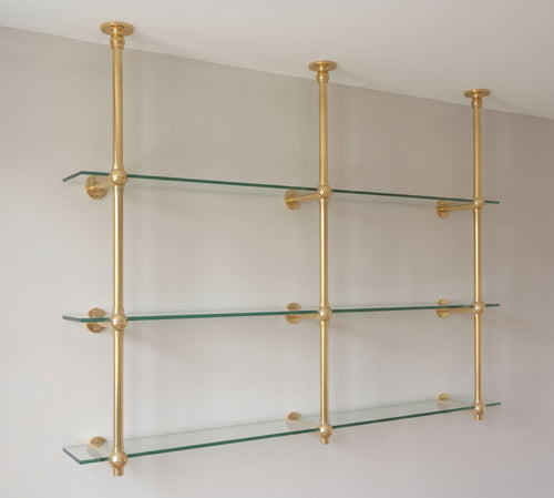 Angled view of the unlacquered brass shelf: three ceiling posts with wall arms holding three tiers of glass