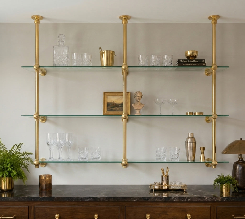Two-bay brass and glass bistro shelf hung from the ceiling, styled with barware above a dark marble sideboard